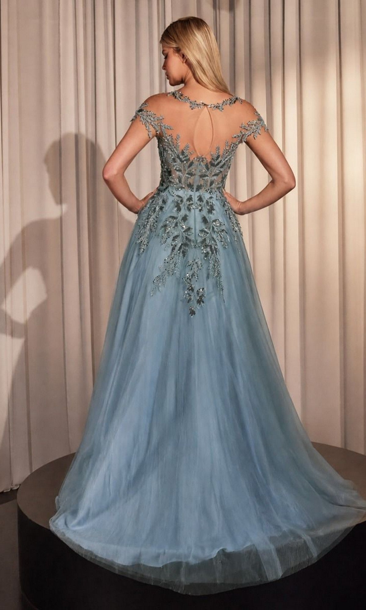 A woman stands on a platform wearing the Andrea and Leo A1475 long prom dress in light blue, featuring floral embroidery, delicate appliques, and a sheer back. Beige curtains hang in the background.